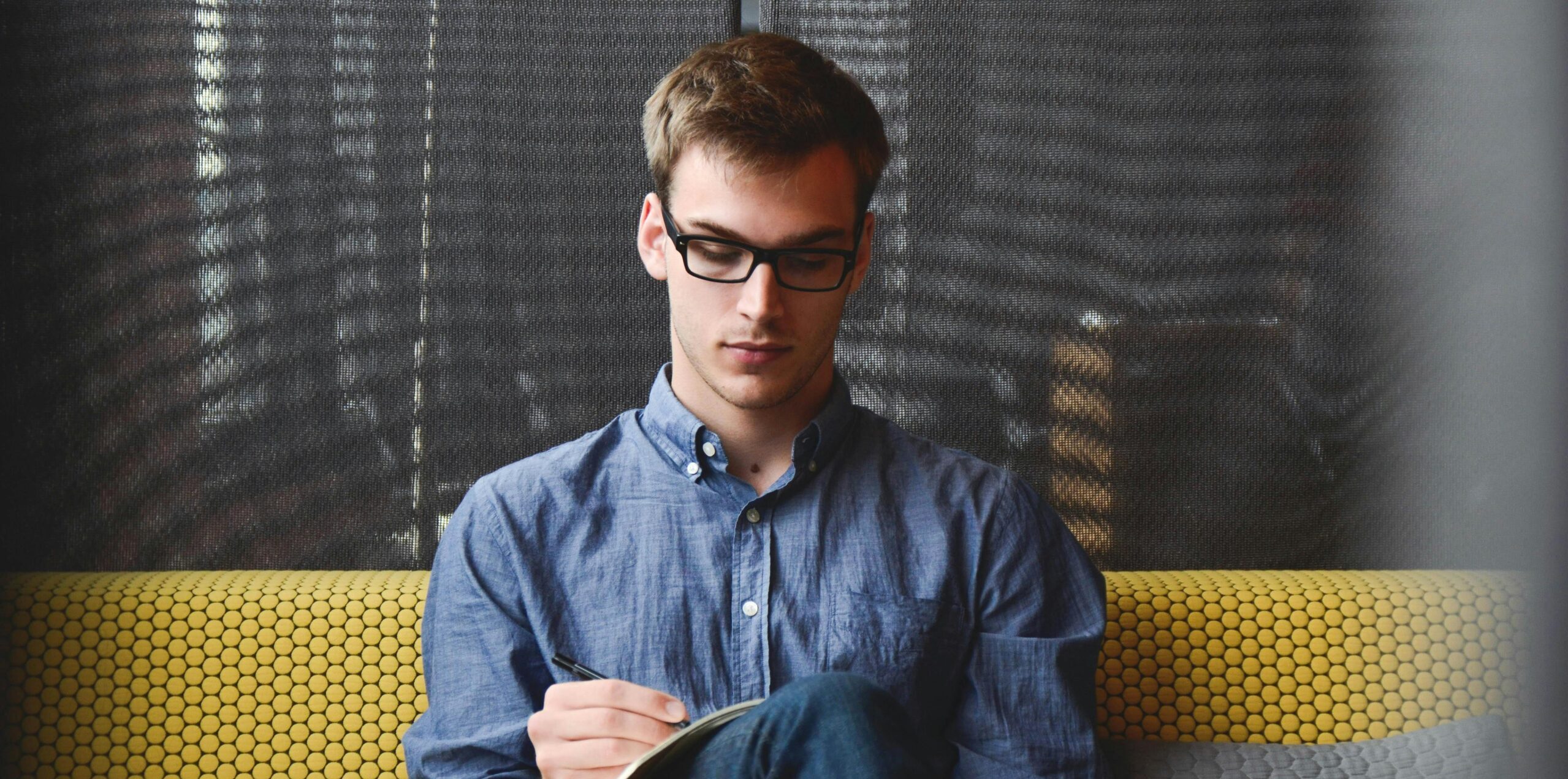A young man in glasses writes in a notebook while sitting on a stylish couch indoors.